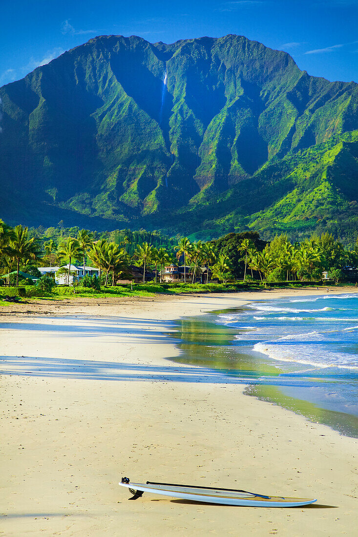 A surfboard sits on the beach at the water's edge with rugged green mountains and lush foliage on the island of Kauai; Hanalei, Kauai, Hawaii, United States of America