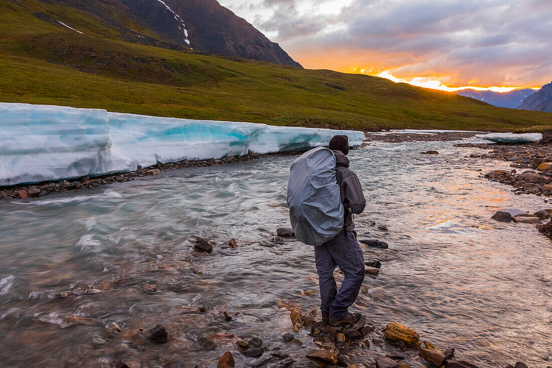 A backpacker observes the midnight sun peeking through the clouds from an unnamed fork of the Atigun River still partially covered in aufeis (sheet-like ice formations) in a remote valley of the Brooks Range; Alaska, United States of America