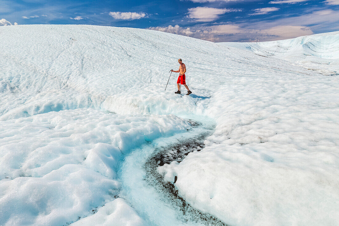 A shirtless man in a bathing suit walks across the surface of Root Glacier with trekking poles in Wrangell-St. Elias National Park; Alaska, United States of America