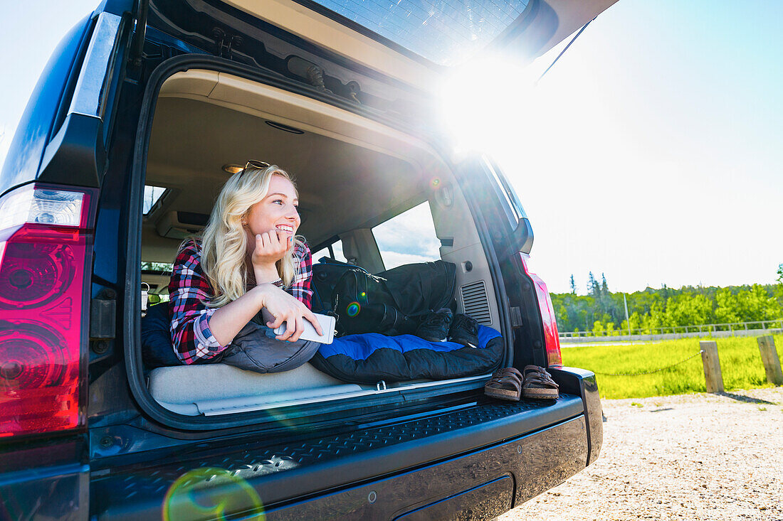A young woman on a road trip lays in the back of a vehicle with her cell phone on a sleeping bag looking out from the  open door; Edmonton, Alberta, Canada