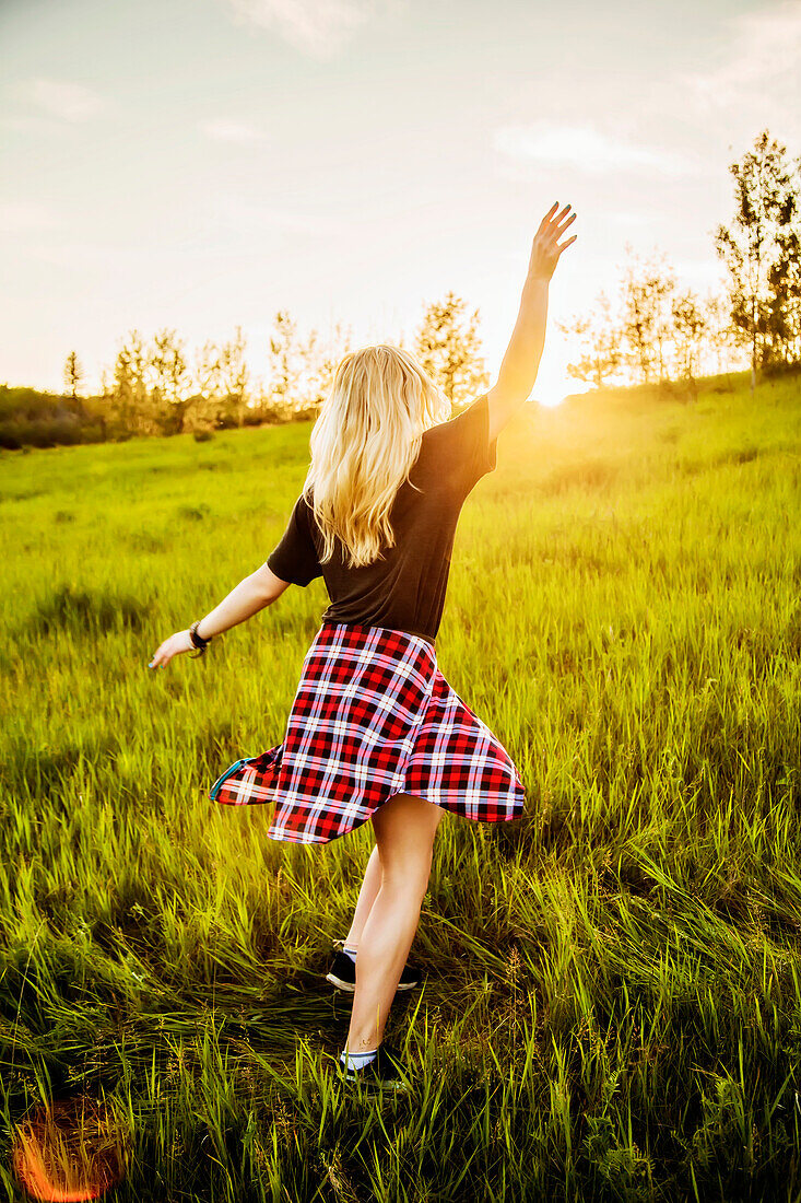 A young woman with long blond hair twirls and runs freely in a grass field in a park at sunset; Edmonton, Alberta, Canada