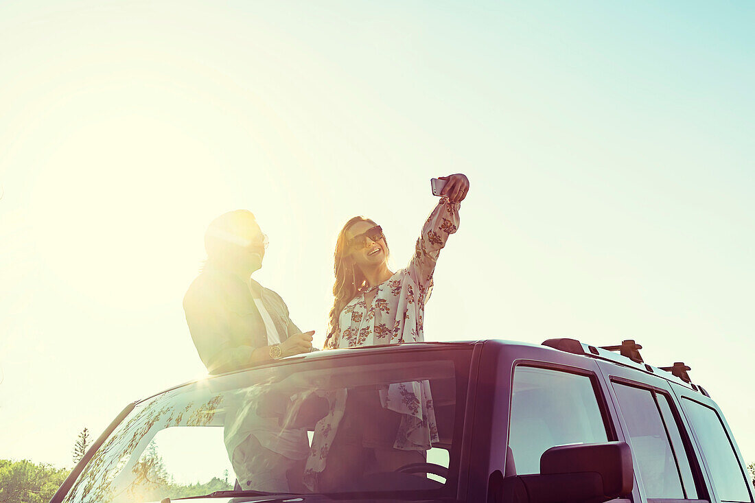 Young couple taking self-portraits with a cell phone while standing up in their sunroof of their vehicle; Edmonton, Alberta, Canada