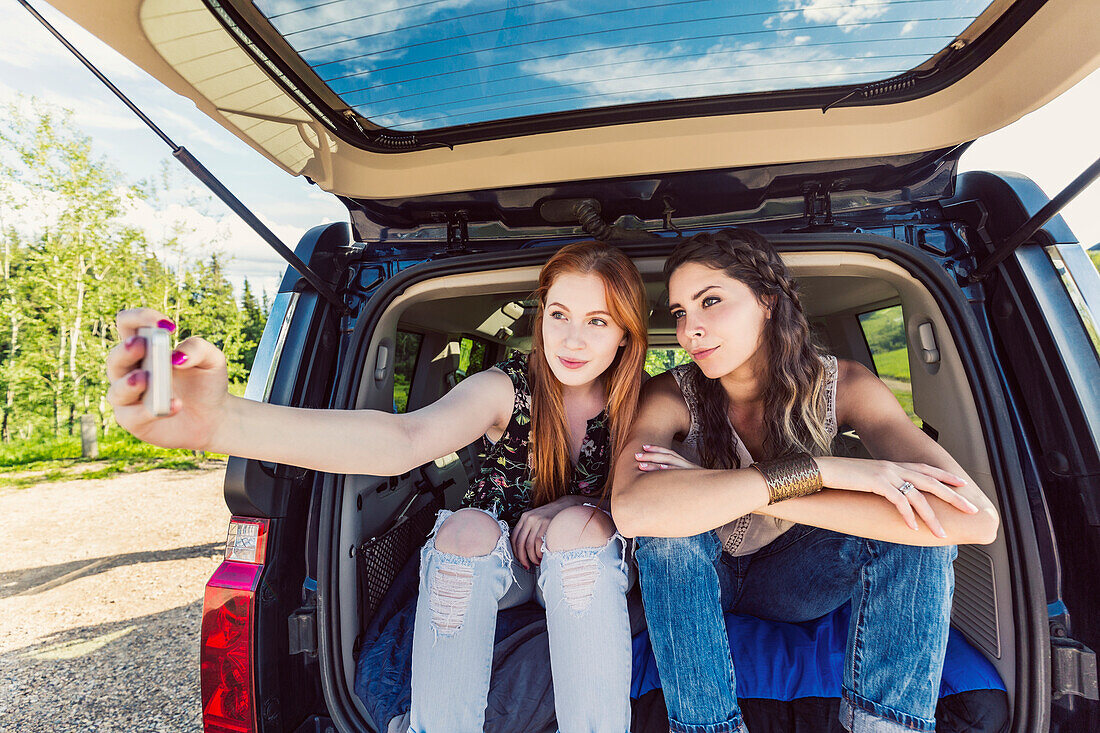 Two young women on a road trip sit in the back of a vehicle taking a self-portrait with a smart phone; Edmonton, Alberta, Canada