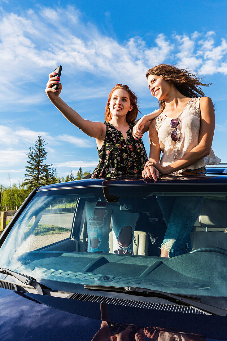 Two young women on a road trip stand up in the sunroof of a vehicle taking a self-portrait with a smart phone; Edmonton, Alberta, Canada