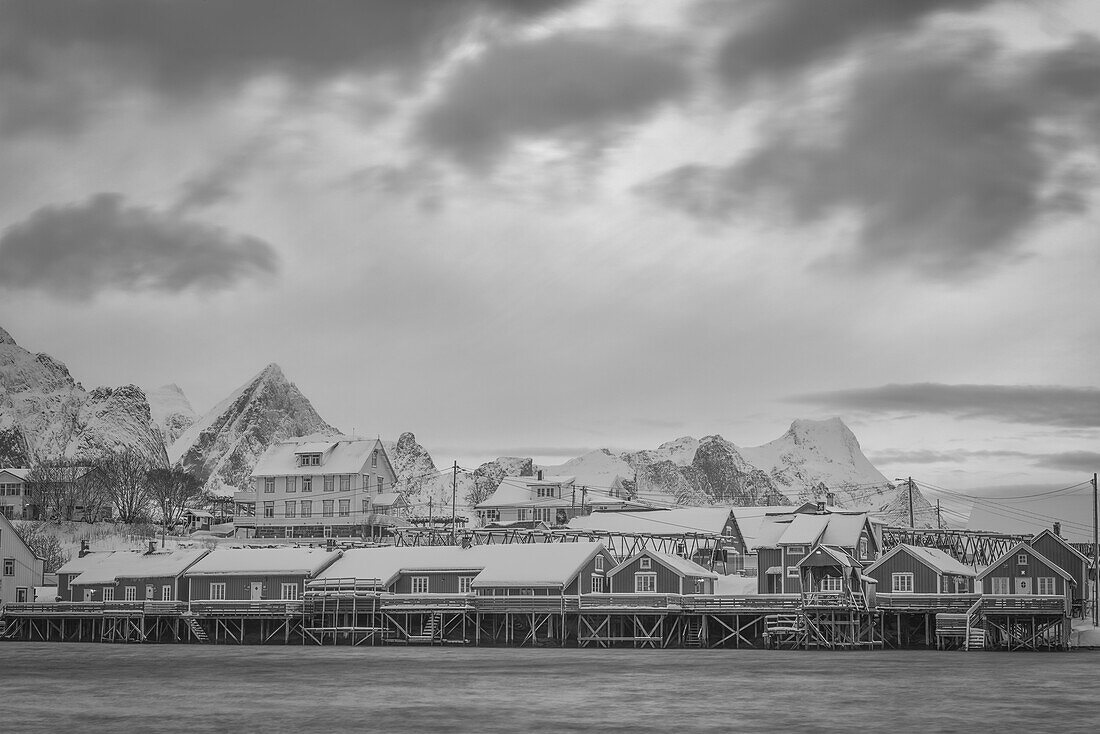 Houses elevated on stilts along the water's edge with rugged mountains in the background and cloud; Svolvar, Lofoten Islands, Norland, Norway