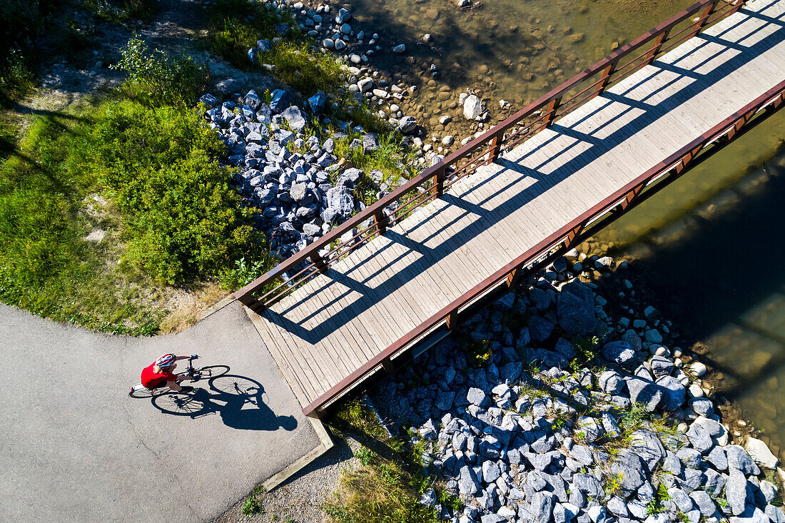 View from directly above a cyclist in a red shirt going down a path and onto a bridge over a creek; Calgary, Alberta, Canada