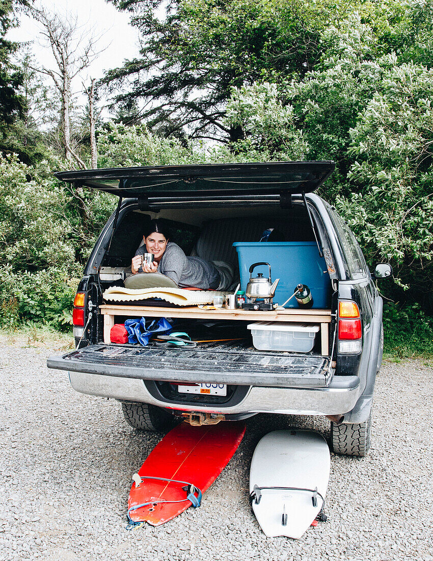 Woman lying in bed in trunk of SUV during surfing road trip, La Push, Washington State, USA