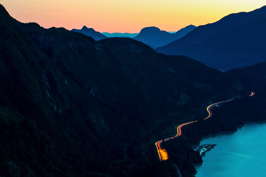 Vehicle light trails on highway stretching between fjord edge and hills at dusk, Squamish, British Columbia, Canada