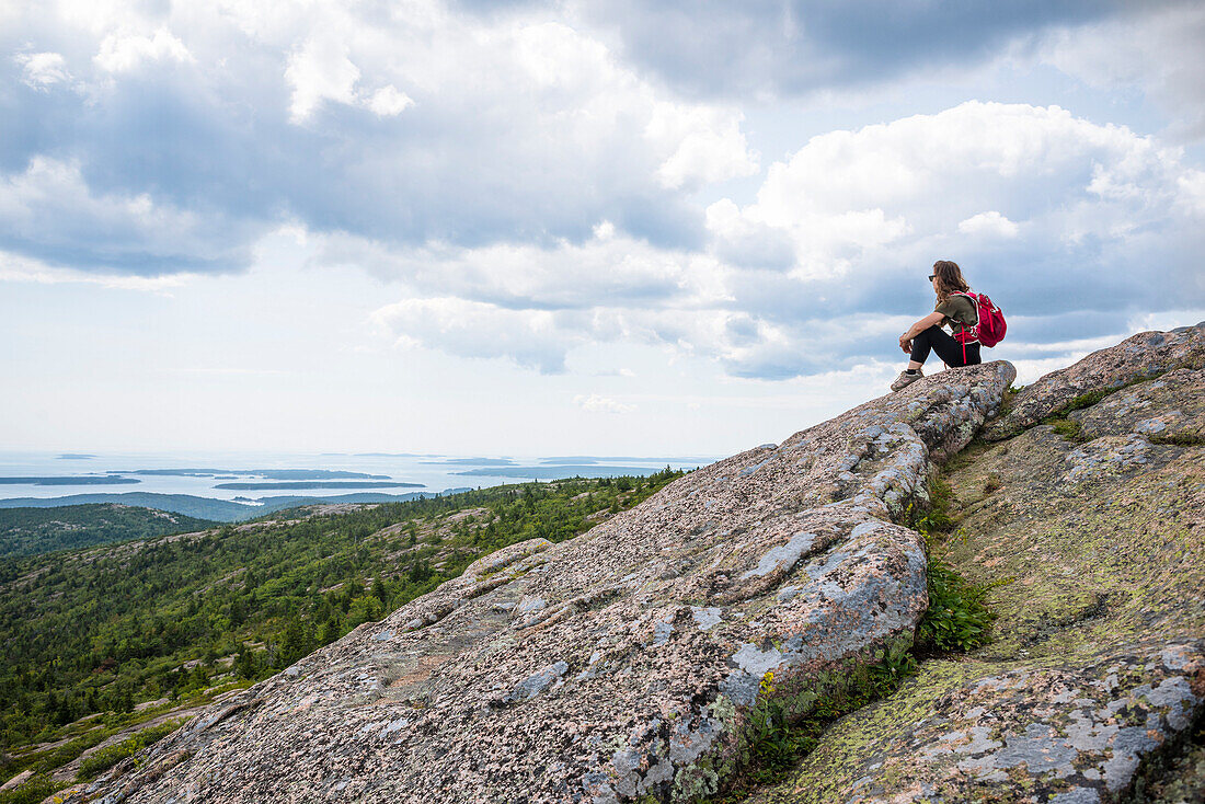 Female backpacker sitting and looking at view from summit of Cadillac Mountain in Acadia National Park, Bar Harbor, Maine, USA