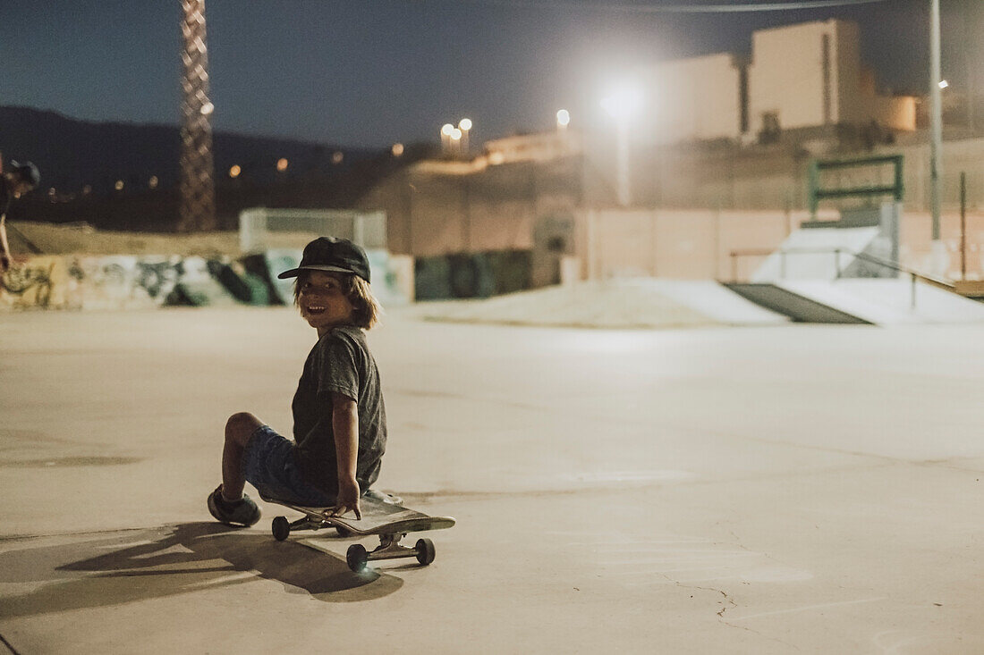 Young skateboarder sitting on skateboard in skate park at night, Tenerife, Canary Islands, Spain