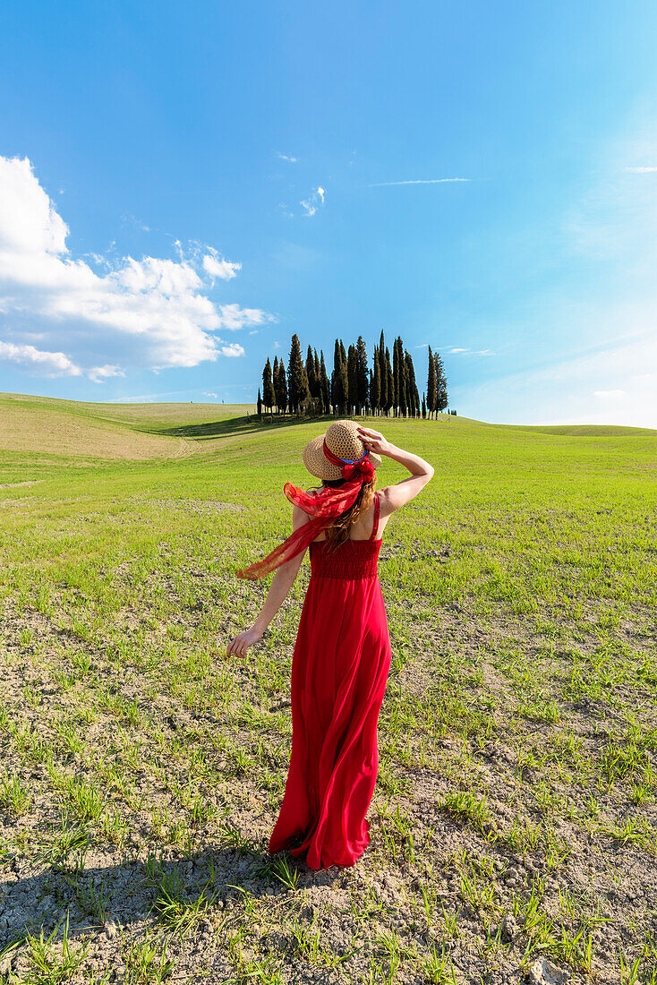 Eine junge Frau im roten Kleid bewundert die Aussicht in einem Weizenfeld in der Nähe der Zypressen des Orcia-Tals