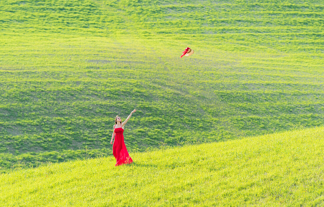 San Quirico d'Orcia, Orcia valley, Siena, Tuscany, Italy. A young woman in red dress is throwing her hat in a wheat field