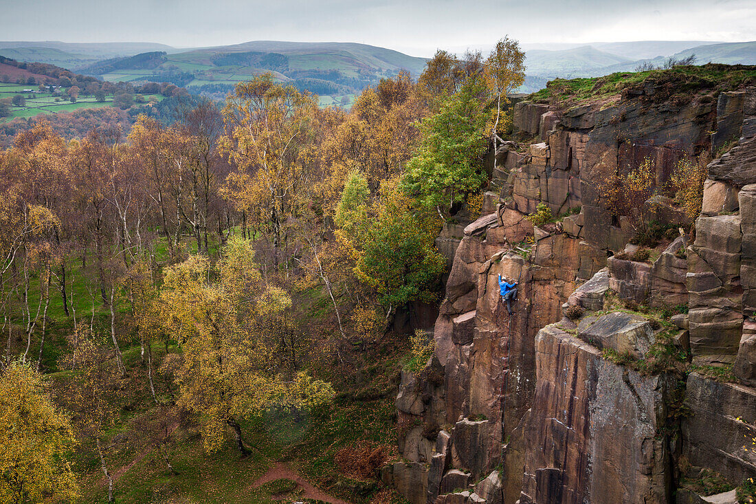 A rock climber ascends a cliff face formed by historic quarrying at Bole hill quarry on an autumn day in the Peak District, Derbyshire, England, United Kingdom, Europe