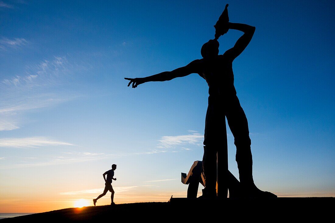 Las Palmas, Gran Canaria, Canary Islands, Spain. A jooger stretches in pre dawn light near sculpture of Greek God, Triton ( son of Poseidon and Amphitrite ) overlooking La Laja beach on the southern outskirts of Las Palmas city.