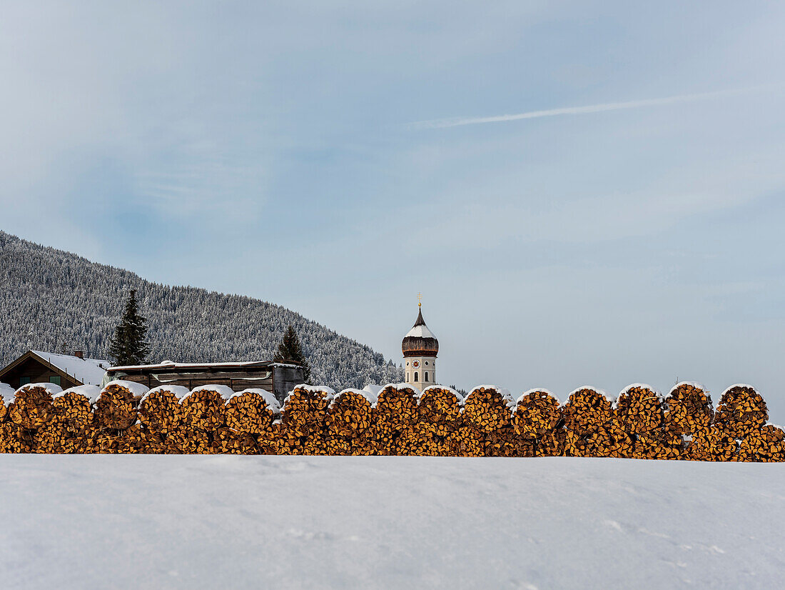 Blick auf schneebedeckte Holzstapel und St. Nikolaus, Unterammergau, Oberbayern, Deutschland