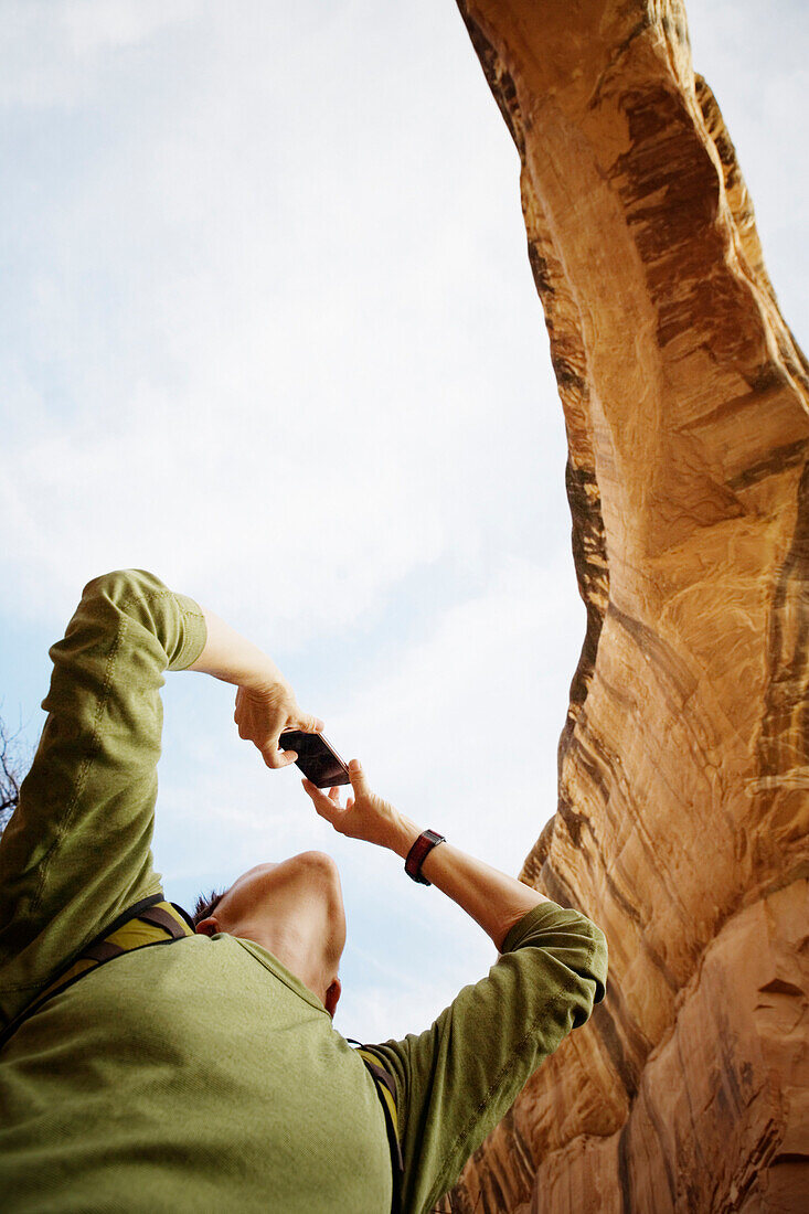 Low angle view of a female hiker taking a photograph with a digital camera under a rock arch in the Natural Bridges National Monument, Utah.