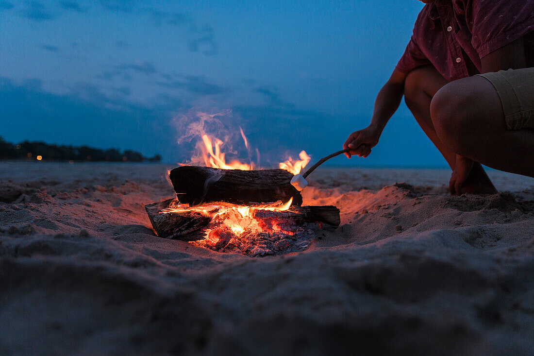 Teenager roasting marsh mellow on beach while kneeling next to small fire in sand, Sheboygan, Wisconsin, USA