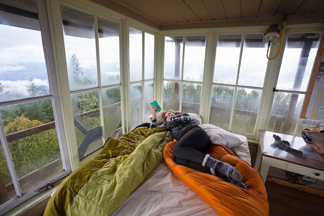 Woman relaxing with phone on bed inside Pickett Butte Fire Lookout near Tiller Oregon, USA