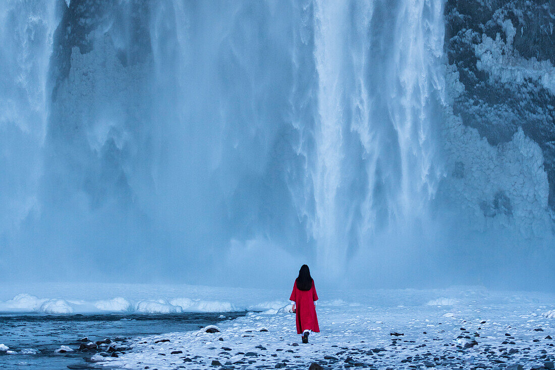 Eine Frau im roten Wintermantel vor dem imposanten Skogafoss Wasserfall, Südküste, Island