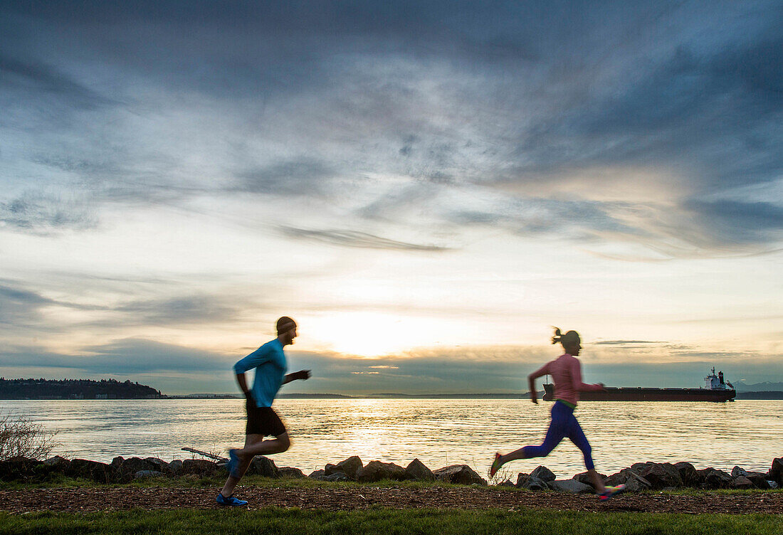 Side view of man and woman running along Puget Sound at sunset, Seattle, Washington State, USA