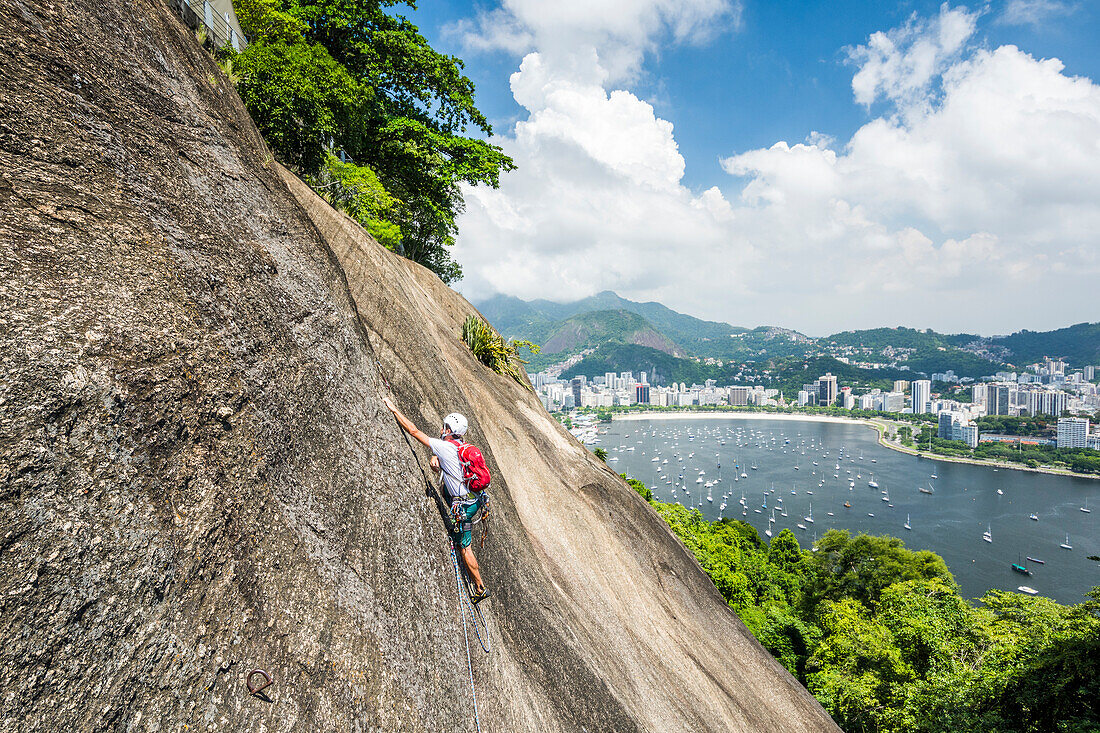 Side view of adventurous man rock climbing Morro da Urca next to the Sugarloaf Mountain with city in background, Rio de Janeiro, Brazil