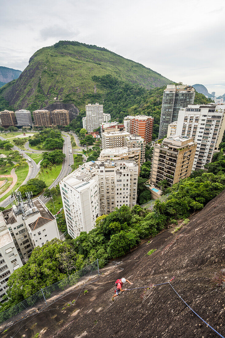 Adventurous man rock climbing Morro do Cantagalo (Cantagalo Mountain) with city in background, Rio de Janeiro, Brazil