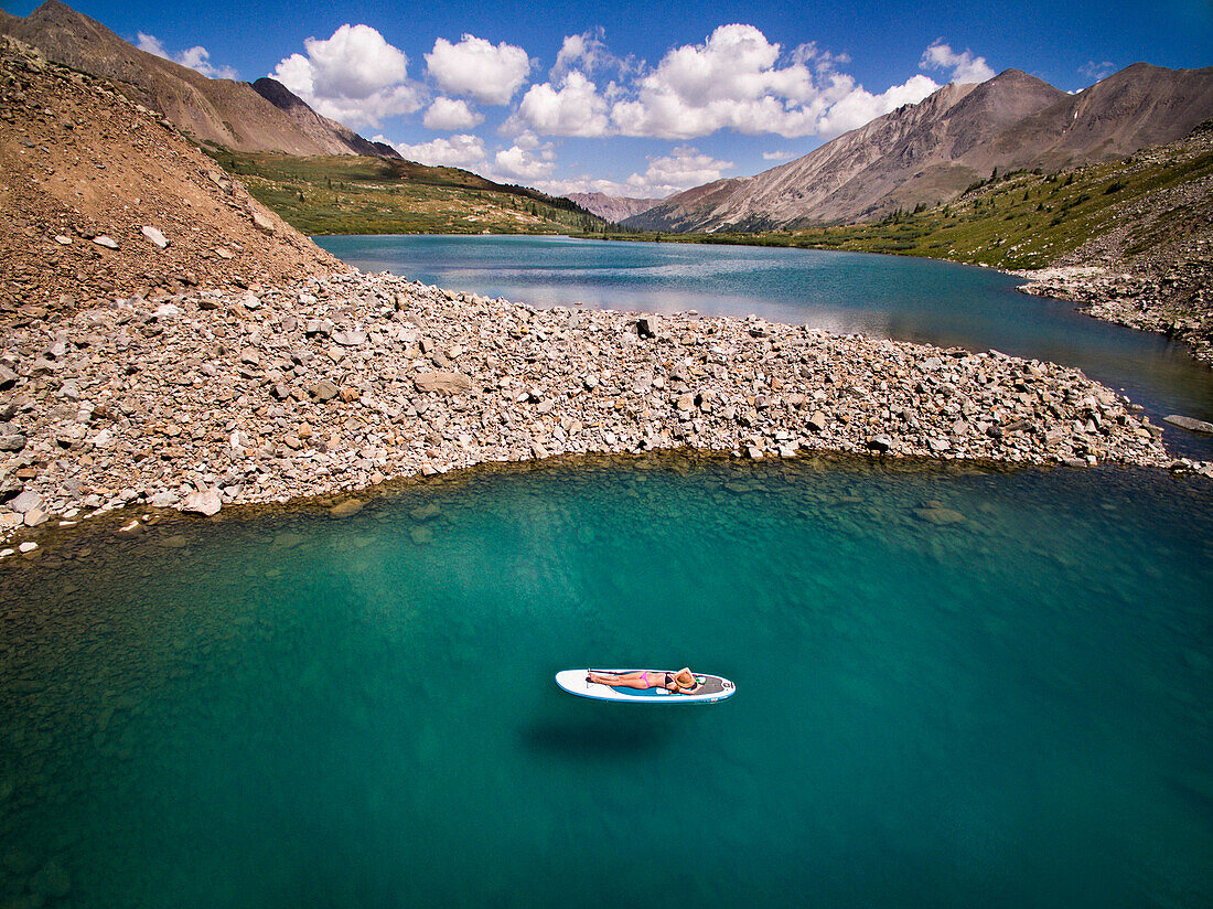 Aerial view of young woman sunbathing while lying on paddle board on high alpine lake in Colorado, USA