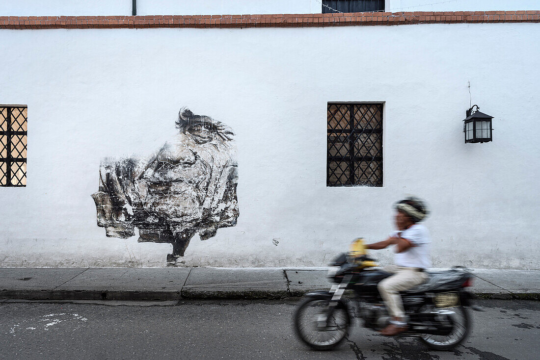 Mann telefoniert mit Handy auf seinem Motorrad während er an einer Wandmalerei eines Gesichts vorbeifährt, Popayan, Departmento de Cauca, Kolumbien, Südamerika