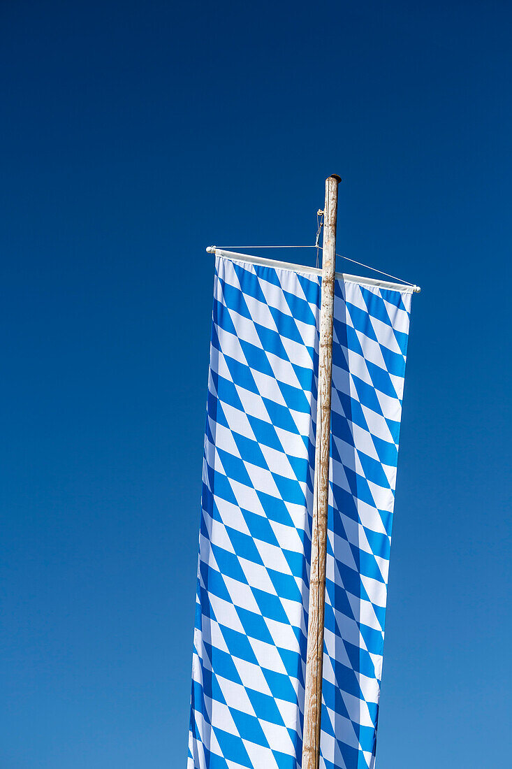 große Bayernfahne mit weiß-blauem Rautenmuster hängt an hölzernem Fahnenmast vor blauem Himmel