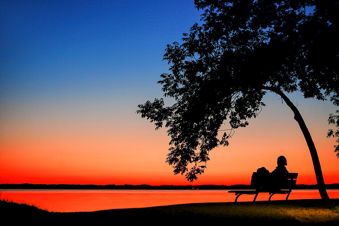 Frau sitzt unter einem Baum auf einer Parkbank am Chiemsee bei farbenprächtigem Sonnenuntergang