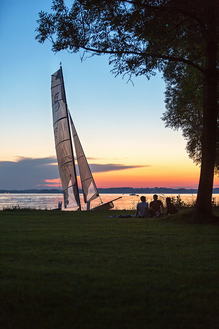 Catamaran is located in the last evening light at the Chiemsee shore, sitting in front of three teenagers under a tree