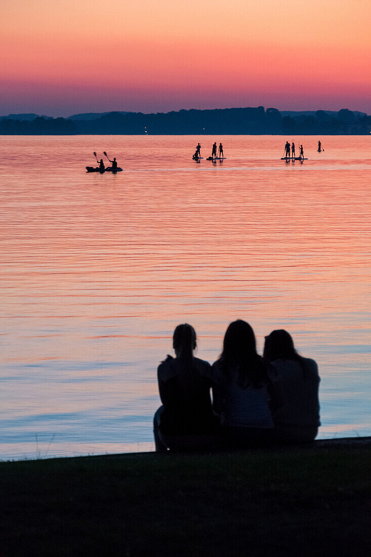 A group of three girls watching standup paddlers and KAYAKERS on the Chiemsee in the sunset