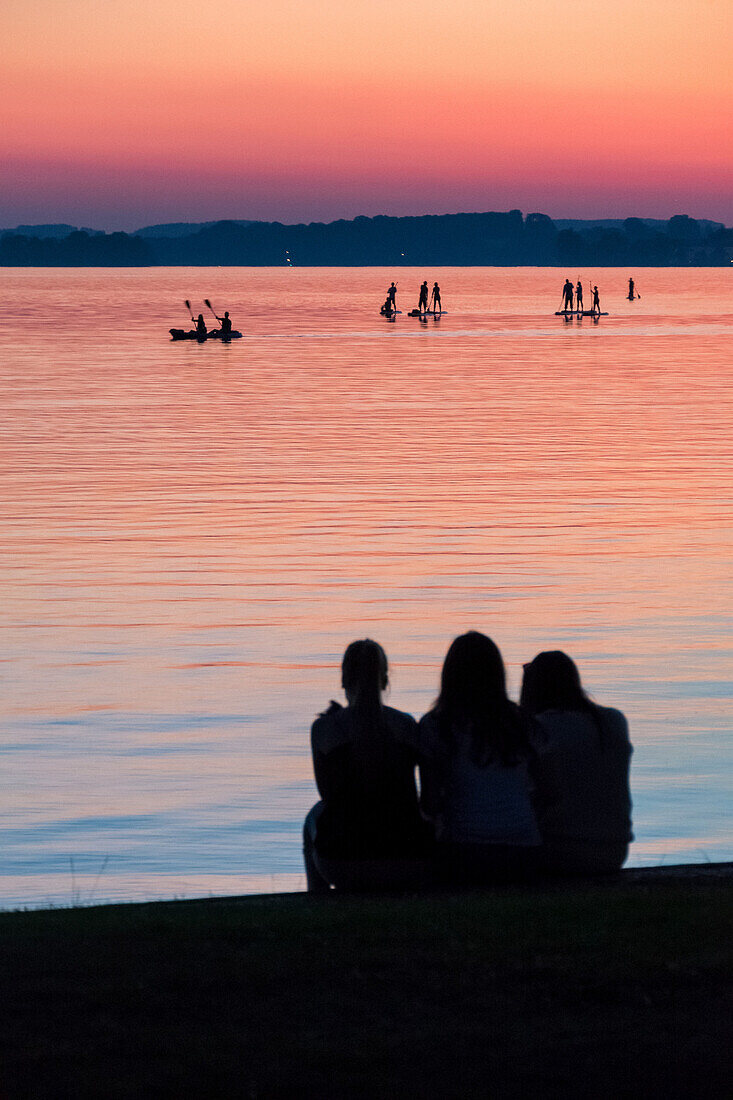drei Mädchen beobachten eine Gruppe von Standup-Paddlern und Kajakfahrer auf dem Chiemsee im Abendrot