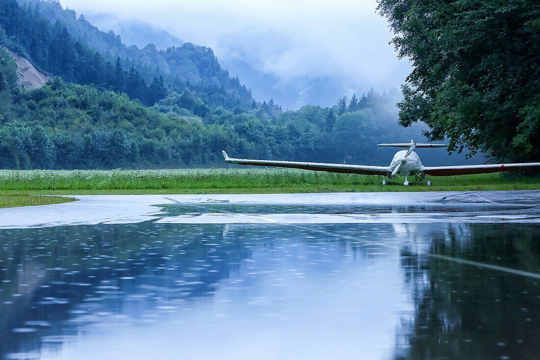 With tarpaulin-covered sports aircraft is located in power rain on the tarmac of the gliding school in unterwössen, forest and mountains in the background