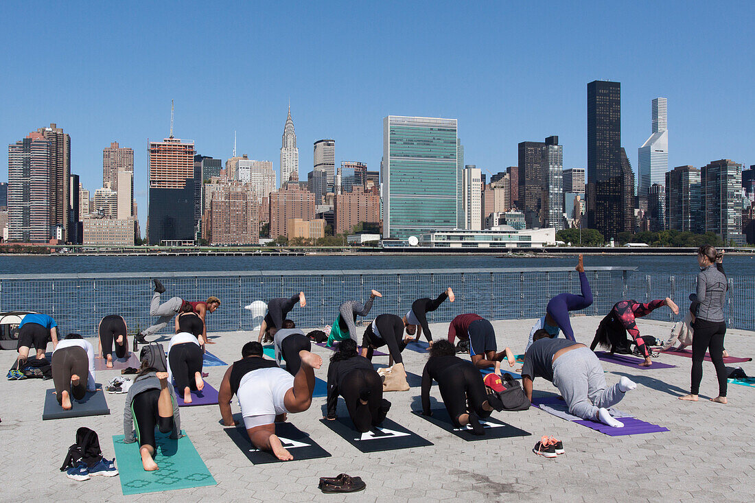 yoga class in gantry plaza state park on the east river across from the skyline of midtown and the headquarters of the un, united nations organization, chrysler building, gantry plaza state park, queens, manhattan, new york city, new york, united states, 