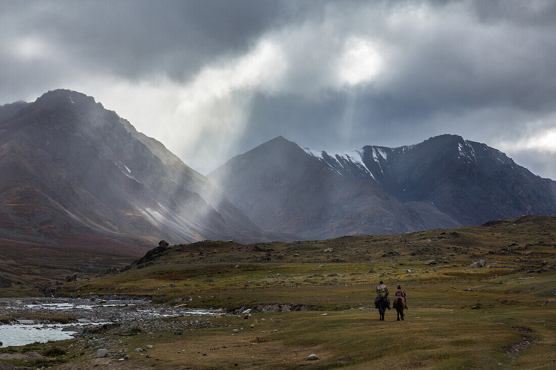kazakh riders beneath the rays of light in cloudy weather, reddish hills with snow-covered summits in the distance, tavan bogd massif, altai, bayan-olgii province, mongolia