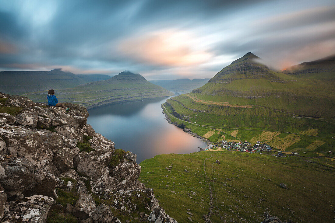 hiker on a promontory contemplating a fjord and a typical village of the faroe islands, funningur, eysturoy, faroe islands, denmark