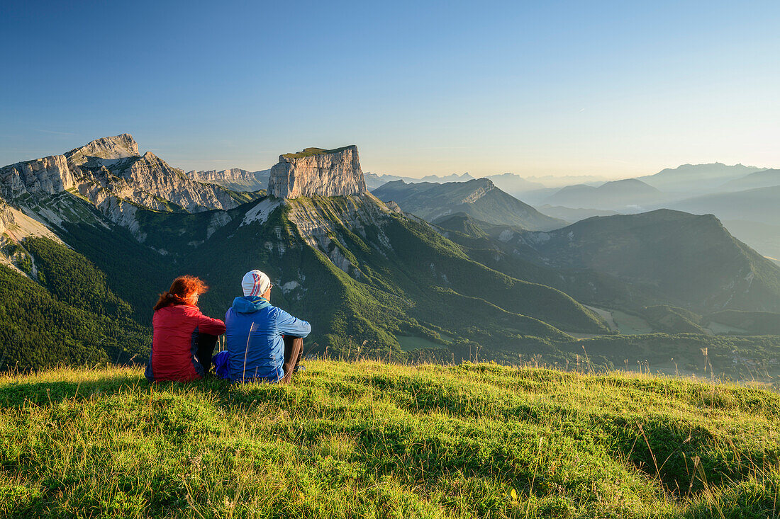 A man and a woman sitting on meadow and views of Grand Veymont and Mont Aiguille in the morning light, from the Tête Chevalier, Vercors, Dauphine, Dauphine, Isère, France