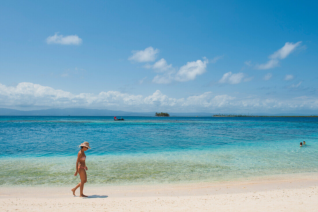 Eine Frau mit Strohhut und Bikini läuft am Strand vor türkisfarbenem Wasser mit einem weit entfernten Boot und einigen Inseln im Hintergrund, San Blas-Inseln, Panama, Mittelamerika