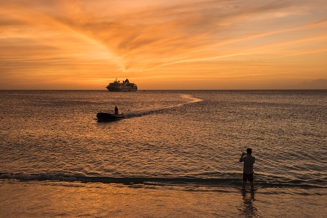 Sonnenuntergangszene mit Expeditions-Kreuzfahrtschiff MS Hanseatic (Hapag-Lloyd Kreuzfahrten) vor Anker am Horizont, ein näherndes Zodiac Schlauchboot und jemand im Vordergrund der ein Foto macht, La Blanquilla-Insel, Venezuela, Karibik