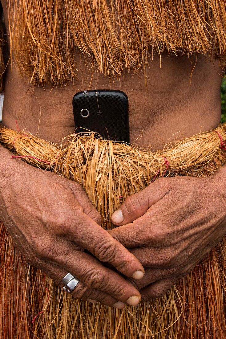 Ein Mitglied des Witoto-Volkes (auch Huitoto) trägt ein Mobiltelefon, das in die Taille seines traditionellen, aus Pflanzenfasern hergestellten Gewand gesteckt ist, Pebas (auch Pevas), nahe Iquitos, Maynas, Peru, Südamerika
