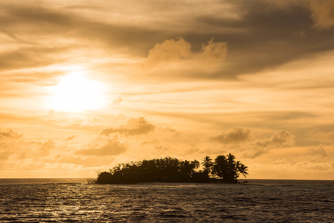 Die Sonne des späten Nachmittags und dünne Wolken malen den Himmel orangefarben hinter einer kleinen Insel, die mit Palmen und anderen Bäumen bedeckt ist, Likiep Atoll, Ratak-Kette, Marshallinseln, Südpazifik
