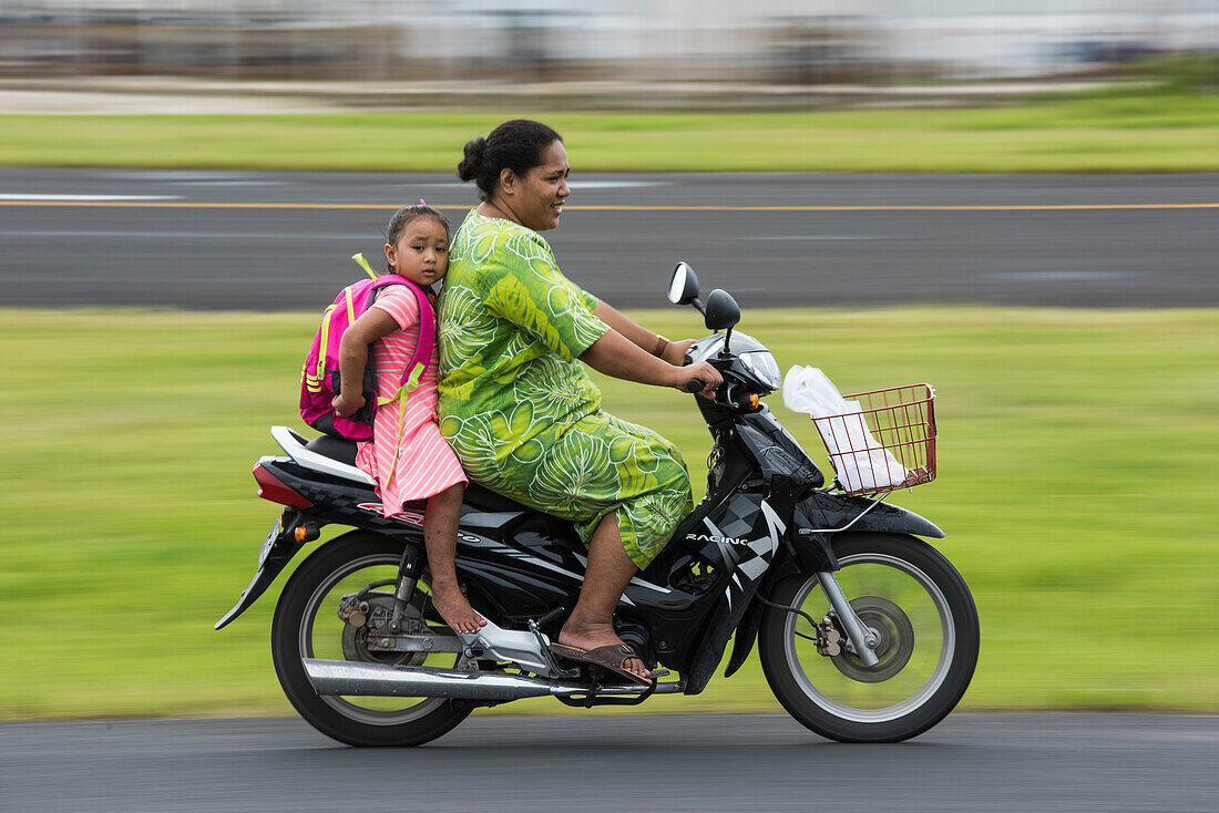 Eine Frau in einem grünen Kleid fährt einen Motorroller mit einem barfüßigen Mädchen in einem rosa-weiß gestreiften Kleid samt Rucksack, Fongafale Island, Funafuti Atoll, Tuvalu, Südpazifik
