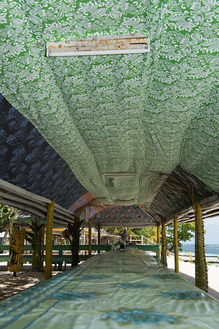 A long table stands under an open-sided roofed structure, decorated with colorful cloths, near a sandy beach, Fagamalo, Savai'i, Samoa, South Pacific