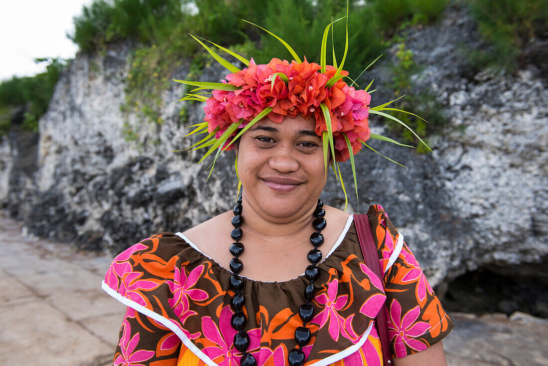 A local woman wearing a brightly colored flower-print dress, a necklace made of large seeds, and a headdress of flowers and leaves smiles into the camera, Atiu, Cook Islands, South Pacific