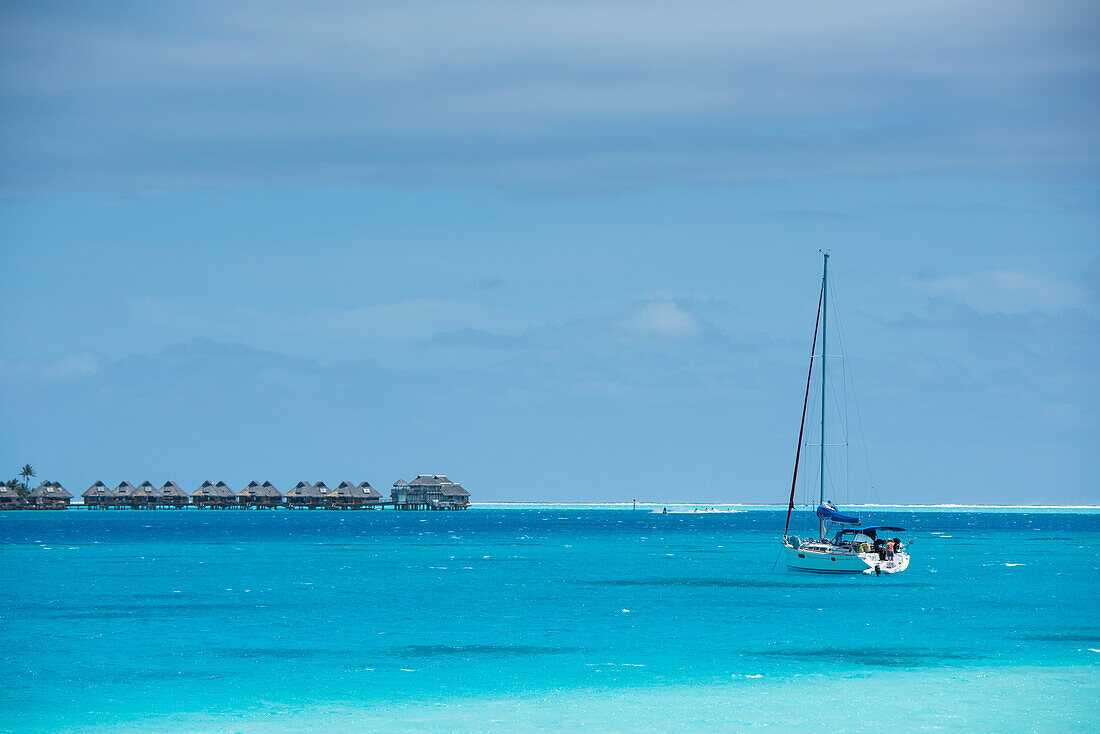 Ein kleines, verankertes Segelboot steht im Vordergrund in türkisfarbenem Wasser, während im Hintergrund eine Reihe von strohgedeckten Überwasser Bungalows eines Luxusresorts auf Stelzen sichtbar ist, Bora Bora, Gesellschaftsinseln, Französisch-Polynesien