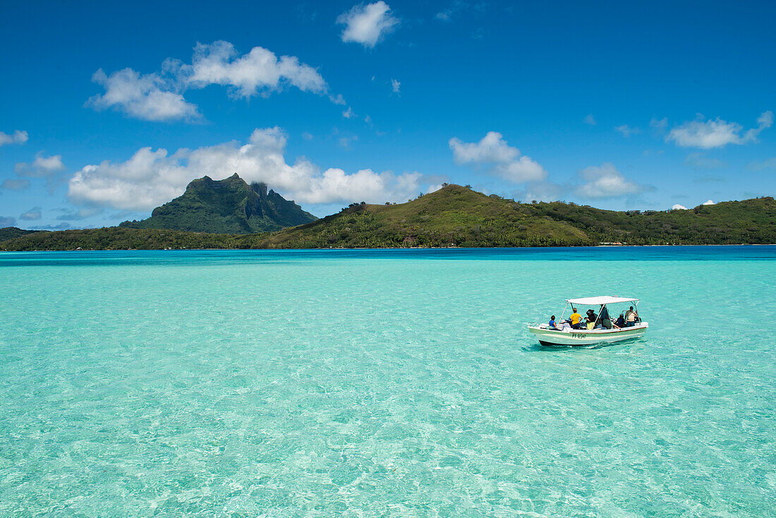 A small motorboat with tourists passes over relatively shallow light-green water in the lagoon, with the island and high mountains in the background, Bora Bora, Society Islands, French Polynesia, South Pacific