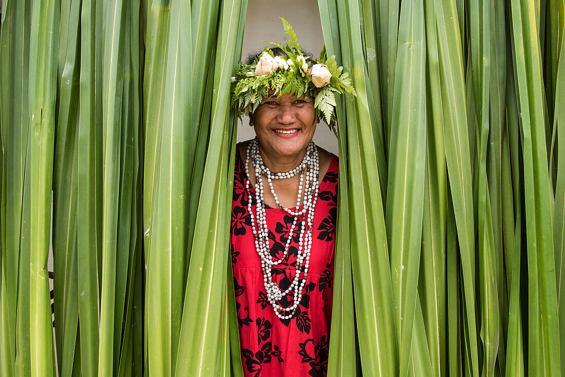Eine einheimische Frau, die ein rotes Kleid mit Blumenmuster, eine Samenkette und einen Kopfschmuck aus Blumen und Farnen trägt, lächelt unter einem Vorhang aus grünen Blättern, Rimatara, Austral-Inseln, Französisch-Polynesien, Südpazifik