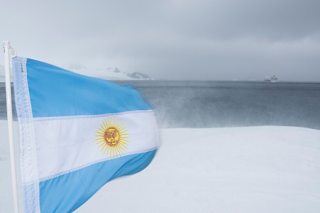 The flag of Argentina blows hectically in heavy blizzard winds at Base Orcadas, while the expedition cruise ship MS Bremen (Hapag-Lloyd Cruises) lies at anchor in the distance, Laurie Island, South Orkney Islands, Antarctica