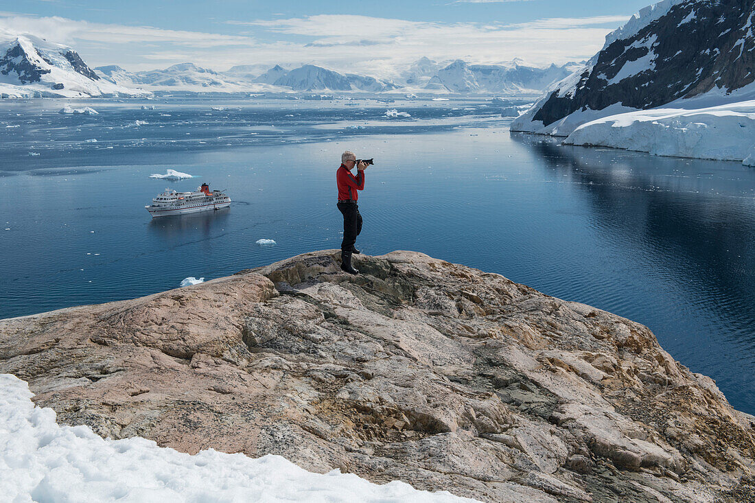 Ein einzelner Fotograf macht Fotos in einer atemberaubend schönen Landschaft umgeben von Bergen, mit Expeditions Kreuzfahrtschiff MS Bremen (Hapag-Lloyd Kreuzfahrten) in der Bucht, Neko Harbor, Graham Land, Antarktis